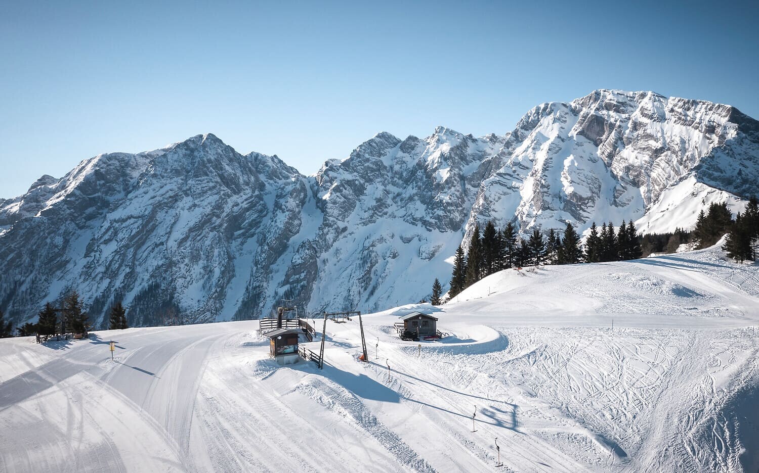 Verschneite Berglandschaft mit Skipiste, zwei Hütten, Schlepplift und Tannen vor schroffen, schneebedeckten Gipfeln.