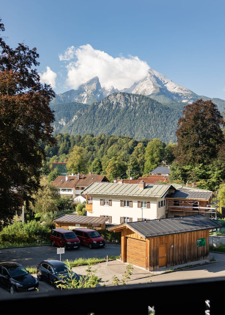 Wolkenverhangener Berggipfel über einem Dorf mit Nadelwald, Häusern und einer Holzhütte sowie geparkten Autos im Vordergrund.