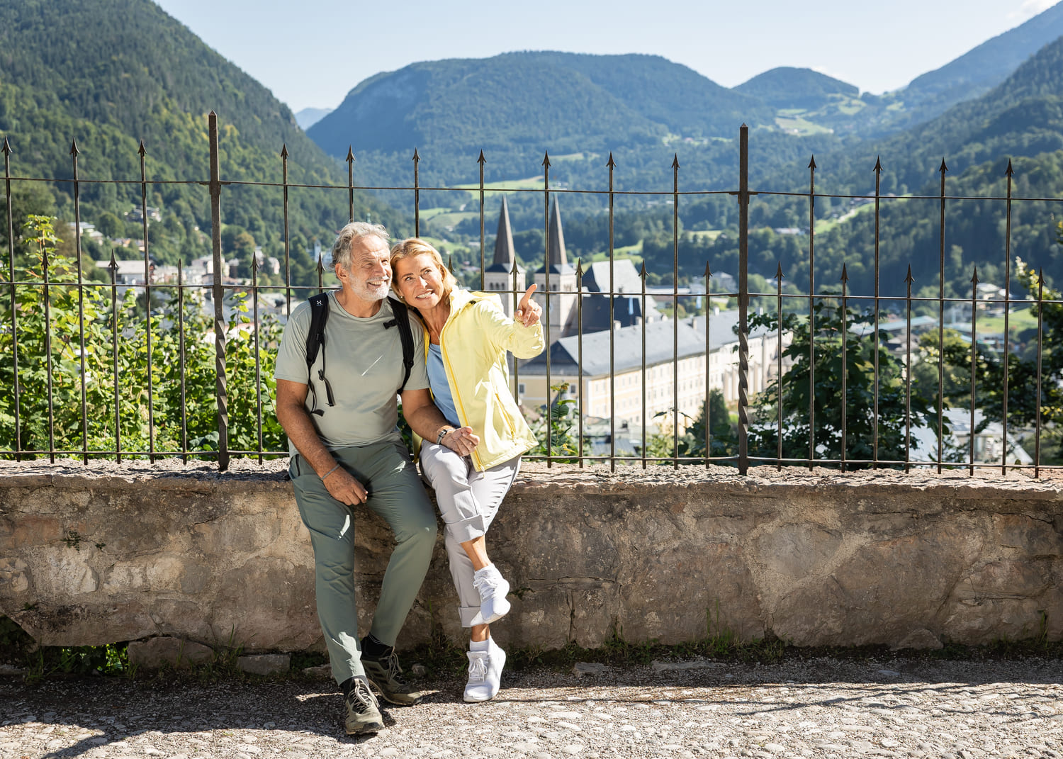 Älteres Paar mit Rucksäcken sitzt auf einer Steinmauer; die Frau zeigt lächelnd auf ein Bergtal mit Kirchturm im Hintergrund.