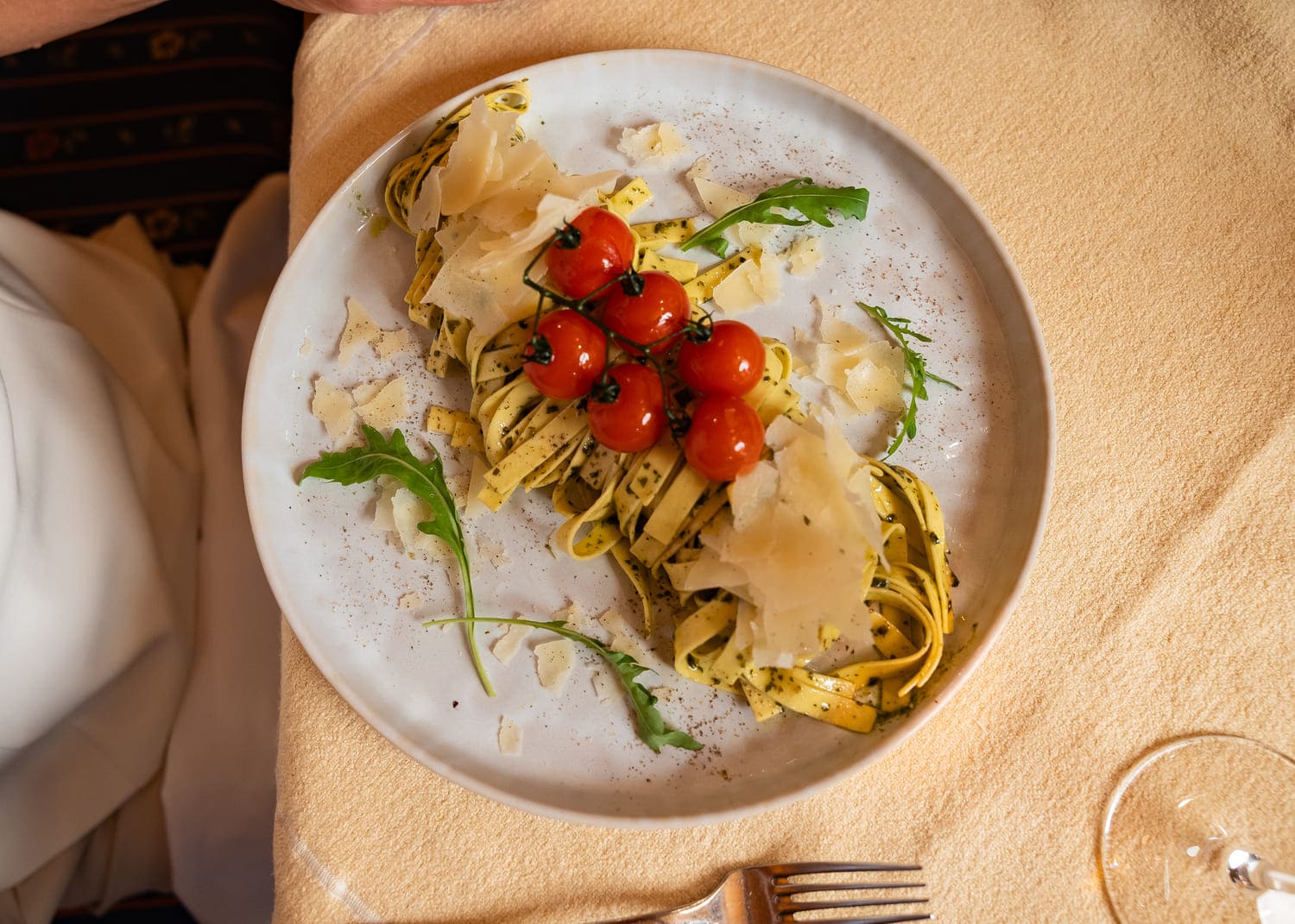 Bandnudeln mit Pesto, Kirschtomatenrispe, Parmesanhobeln und Rucola auf hellem Teller.