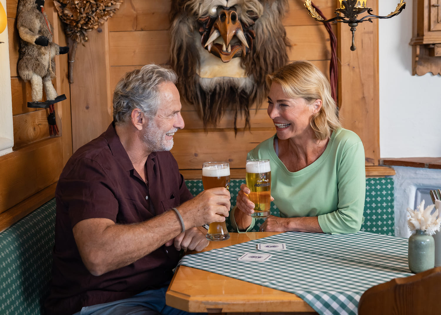 Älteres Paar lacht und stößt mit zwei Biergläsern an in einer gemütlichen Holzstube