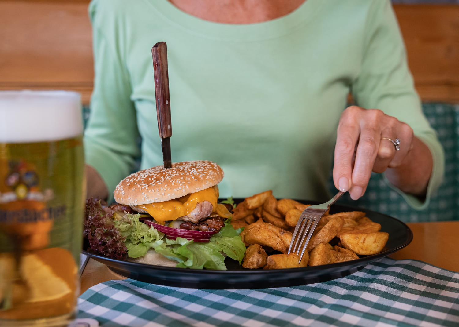 Person in grünem Shirt isst Cheeseburger mit Sesambrötchen und senkrechtem Messer; Gabel sticht Kartoffelspalten, Bier vorn.