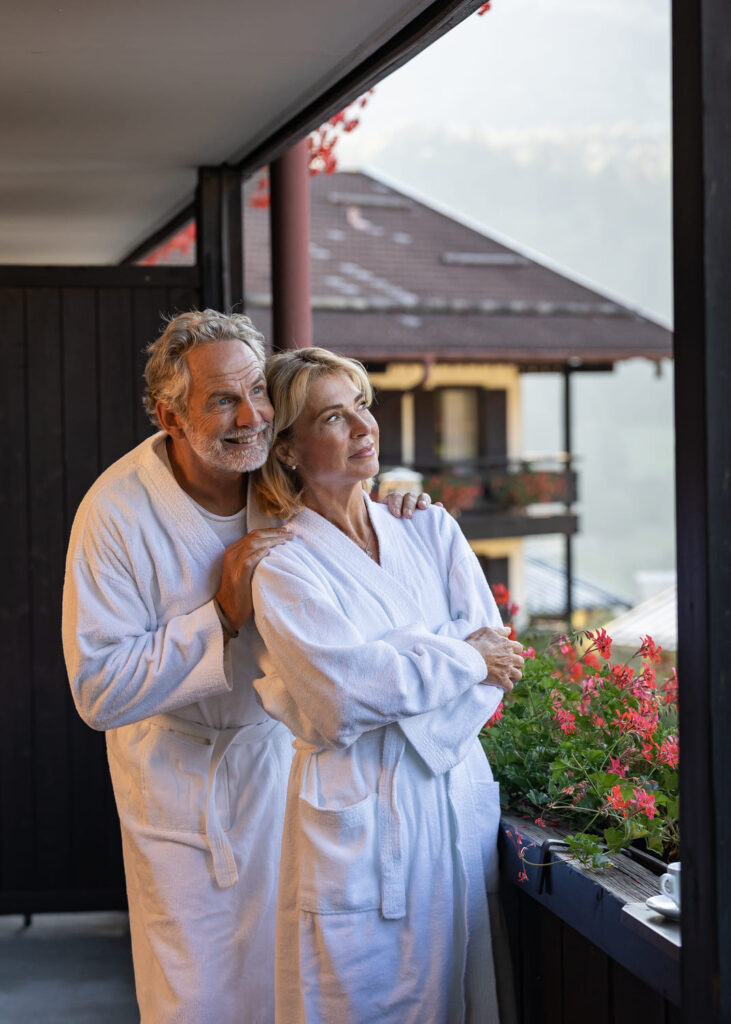 Älteres Paar in weißen Bademänteln auf Balkon, Mann umarmt Frau, beide blicken lächelnd in die Ferne; rote Balkonblumen.